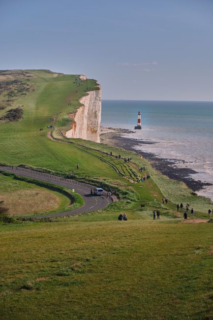 A gravel pathway leading down to a small cluster of white residential houses with dark chimneys situated near the coastline. Behind the houses, there is a sandy beach and calm sea waters. The scene is