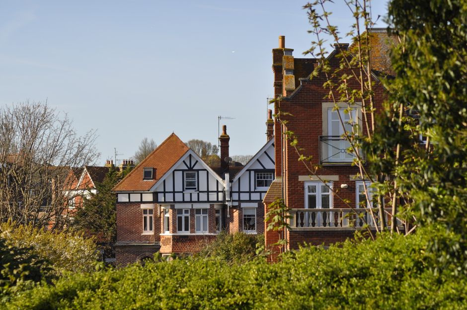 A view of traditional residential buildings with red brick exteriors, white window frames, and decorative timber detailing, set against a clear blue sky. The foreground features lush green foliage and